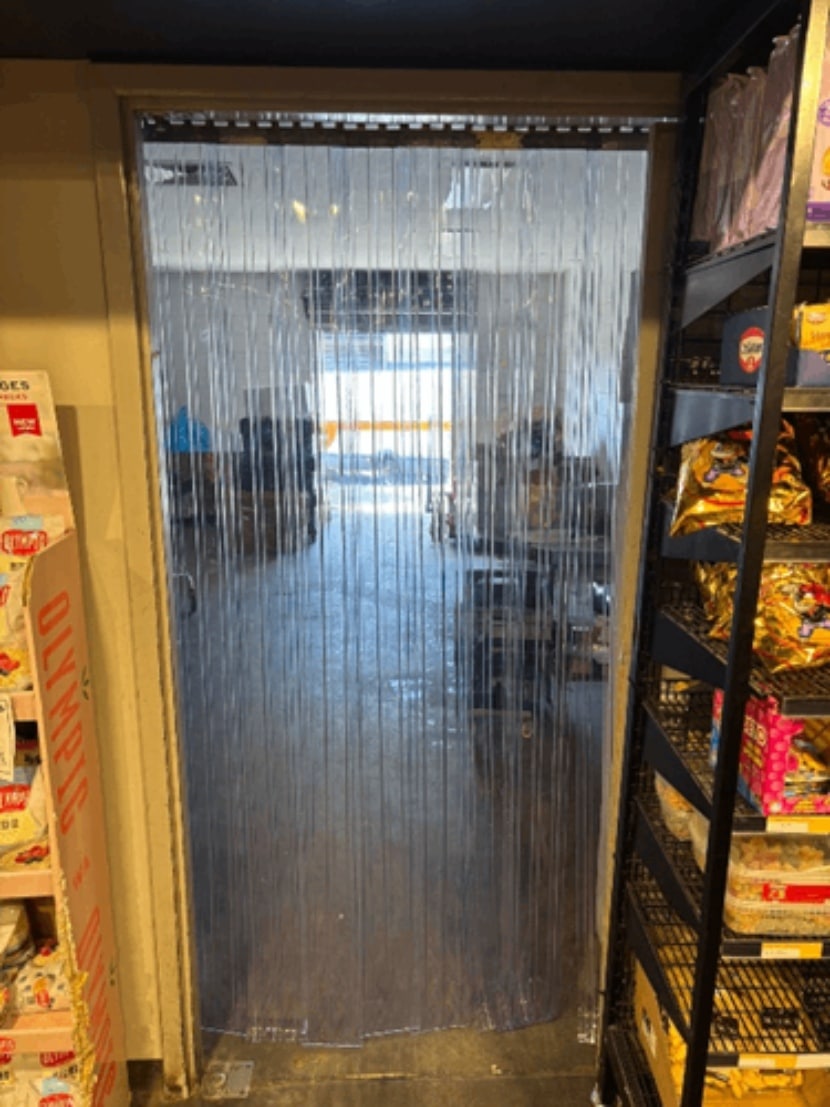 Transparent PVC strip curtain installed in a store doorway, separating retail shelves from a storage area.