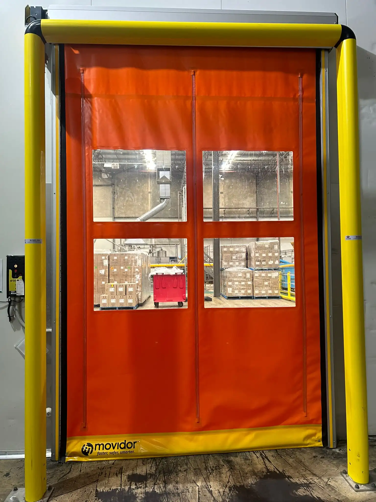 An orange Movidor high-speed roll-up door with four transparent windows, flanked by bright yellow safety bollards in a warehouse setting.
