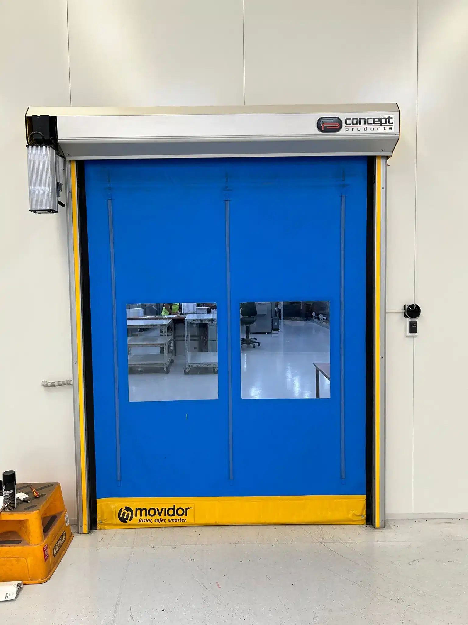 A blue Movidor high-speed roll-up door with two transparent windows, installed indoors in a commercial facility. A motor is mounted on the top left.