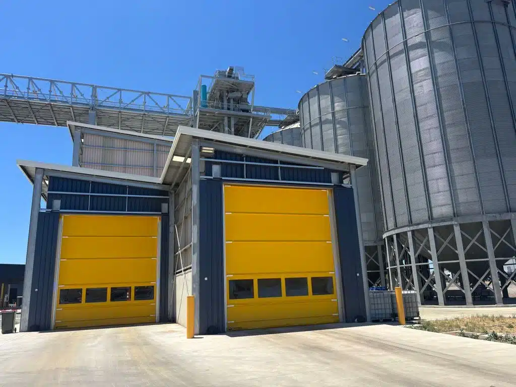 Two bright yellow industrial high-speed doors installed on a grain storage facility, flanked by large corrugated steel silos and overhead conveyors.
