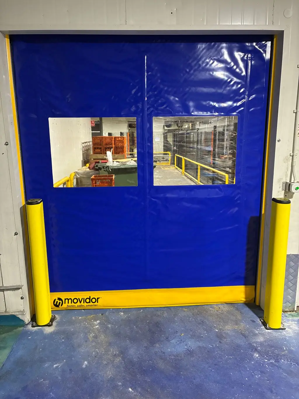 Blue Movidor rapid door with viewing windows, protected by yellow DEFLEX safety posts on each side
