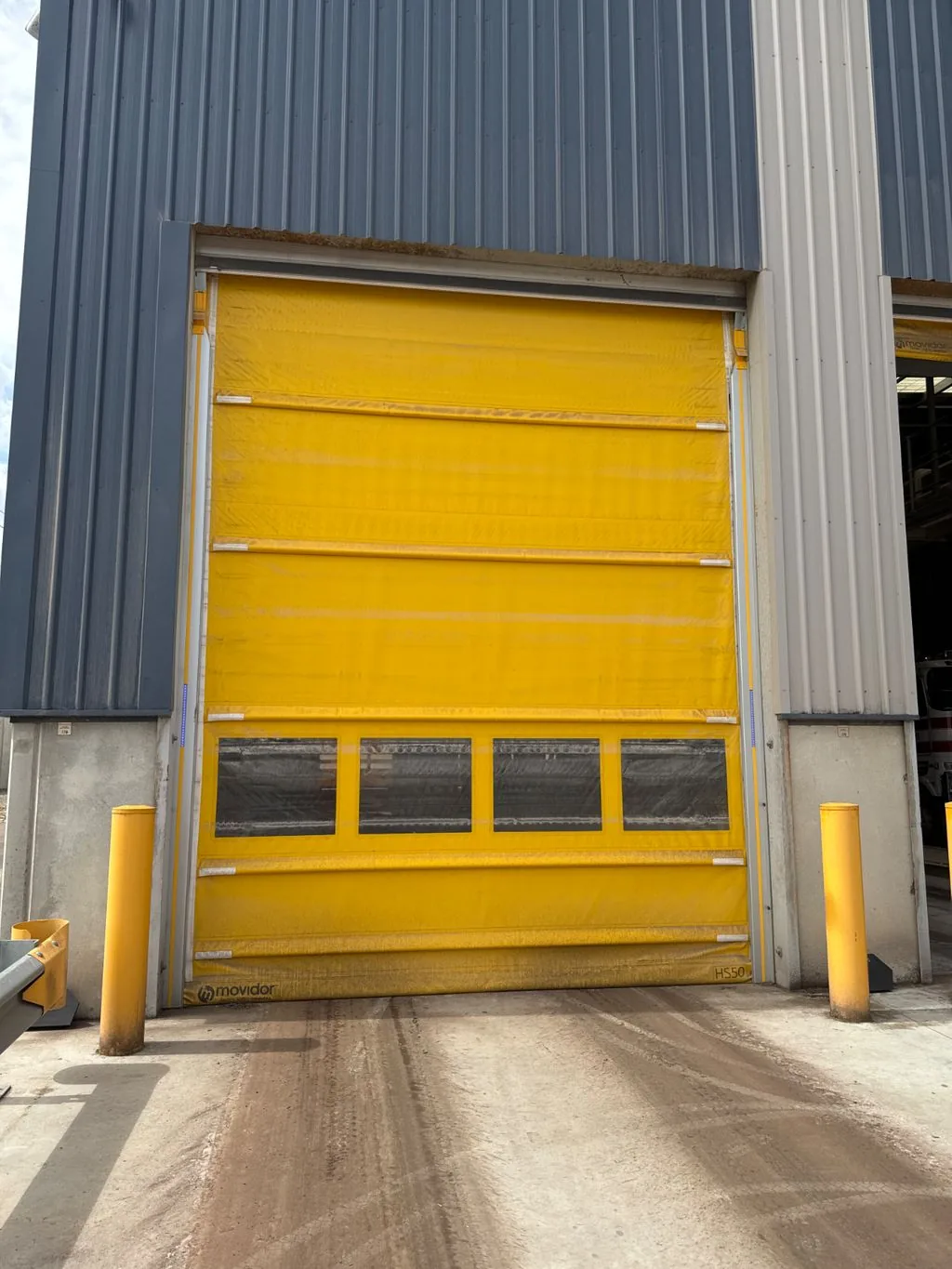 Yellow Movidor HS50 rapid roll door with vision panels, installed in a steel-clad industrial building between yellow safety bollards